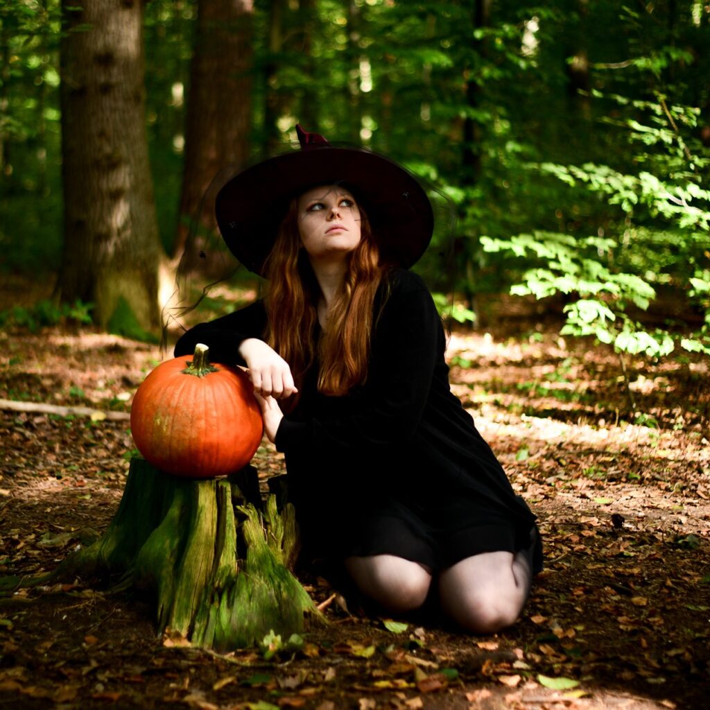 Eine Hexe komplett in schwarz gekleidet, sitzt im Wald und lehnt sich an einem Kürbis an, während ihr Blick in den Himmel gerichtet ist.