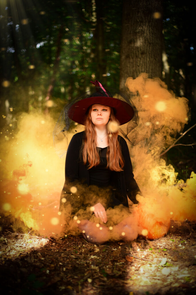 Fantasy-Foto einer düster gekleideten Hexe im Wald, eingehüllt von bedrohlich orangenem Rauch ihres Zauberspruchs.