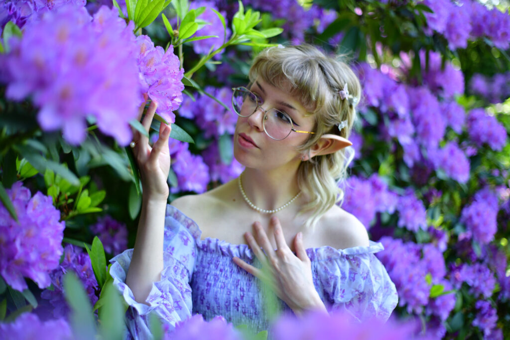 Fantasy-Portrait einer Elfe mit Brille, im lila Kleid und Perlenschmuck vor einem Meer aus lila Blumen.