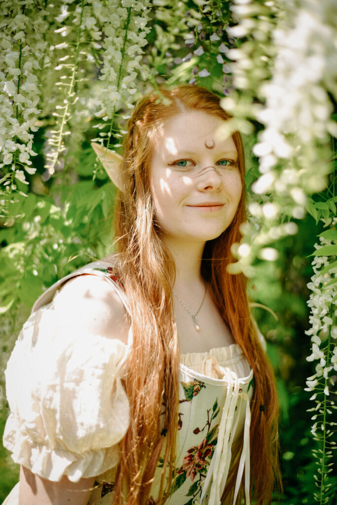 Portrait von Kimi-chan als Elfe im hellen Korsettkleid vor einem Baum mit weißen Blüten.