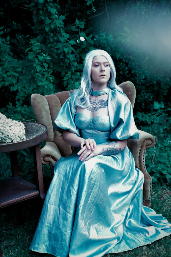 Eine anmutige Frau mit weiß-silber Haar und einem silber-blauem Kleid sitzt auf einem braunem Stoffsessel. Neben ihr ein Tisch mit Blumen drauf.