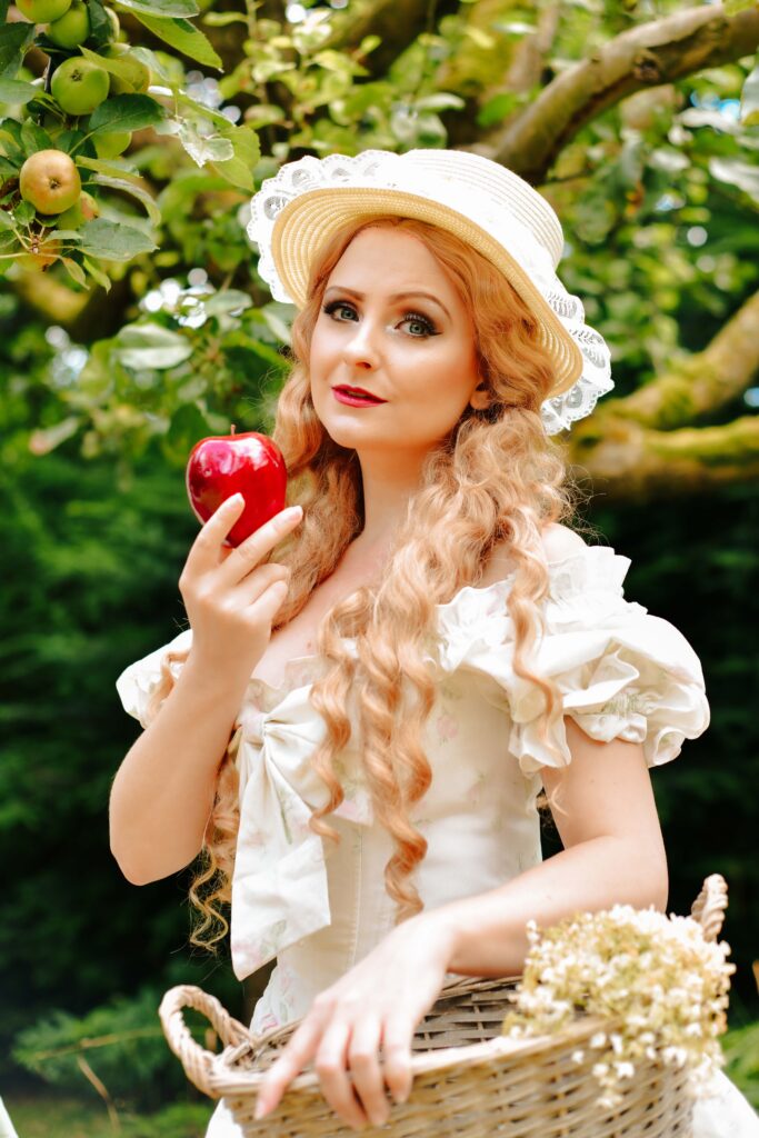Eine junge Frau mit blond rotstichigen Haaren trägt ein weißes Kleid und einen weißen Hut mit Rüschen. Sie trägt einen Korb mit Blumen und hält einen roten Apfel in der Hand.