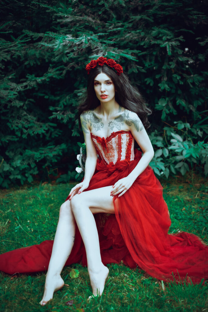 Eine dunkelhaarige Frau mit bleicher Haut trägt einen rote-Rosen-Kopfschmuck und ein rotes Korsagen-Kleid mit Rüschen. Sie sitzt auf einem Stuhl.