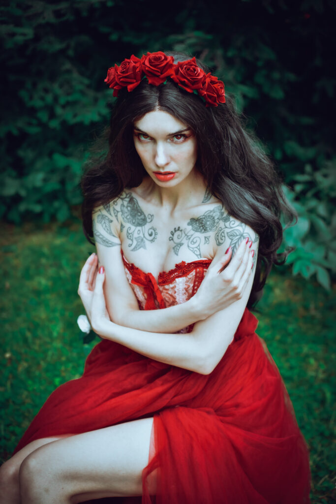 Eine dünne Frau mit weißer Haut, dunklen Haaren, einem roten Kleid mit rotem Kopfschmuck aus Rosen sitzt draußen, umgeben von dunklen Tannen.