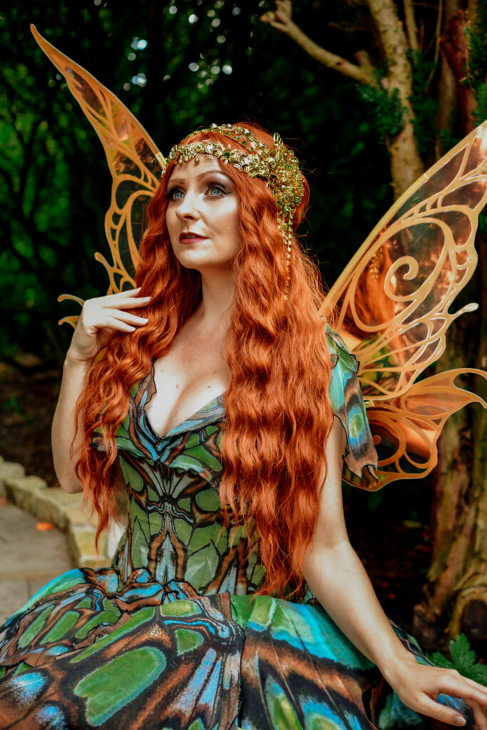 Ein Elfen-Portrait aus meinem Elfen-Fantasy-Fotoshooting. Sie trägt goldenen Kopfschmuck, ein Kleid mit grün braunem Mottendesign, hat orange goldene Flügel und rote Haare.