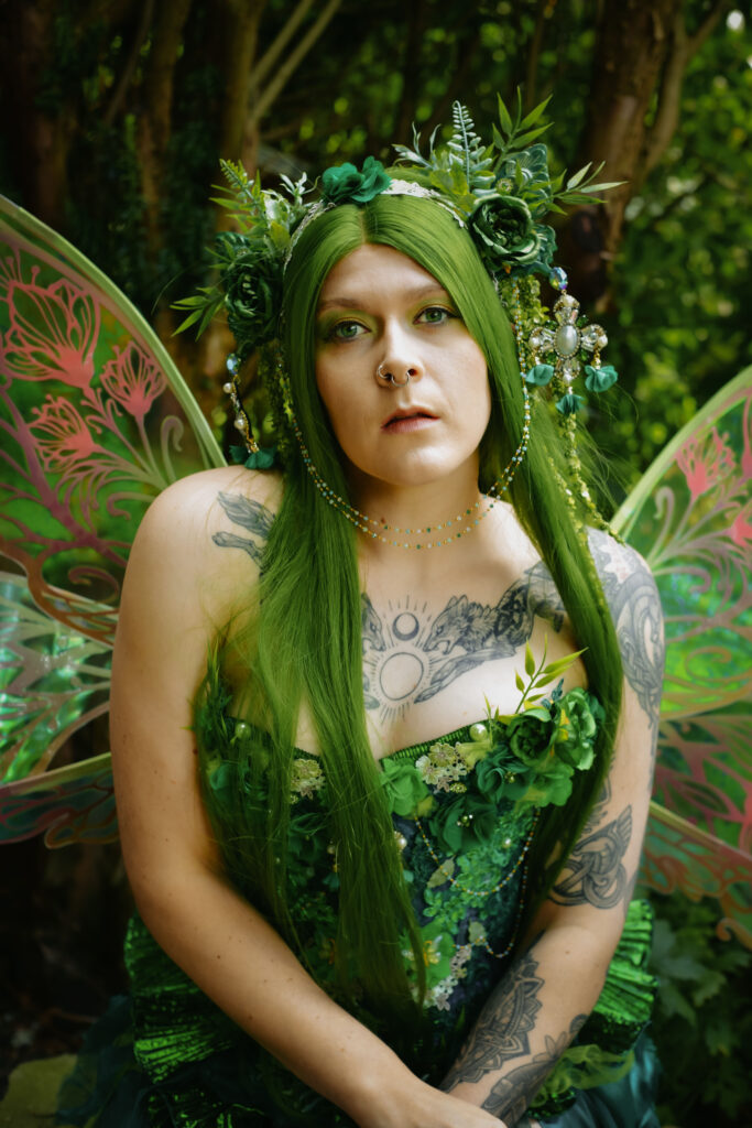 Eine Waldelfe mit einem grünem Kleid, auf dem Pflanzen wachsen sitzt im Wald auf einer Mauer. Sie hat grüne Flügel mit ebenfalls mit Blumenelementen.