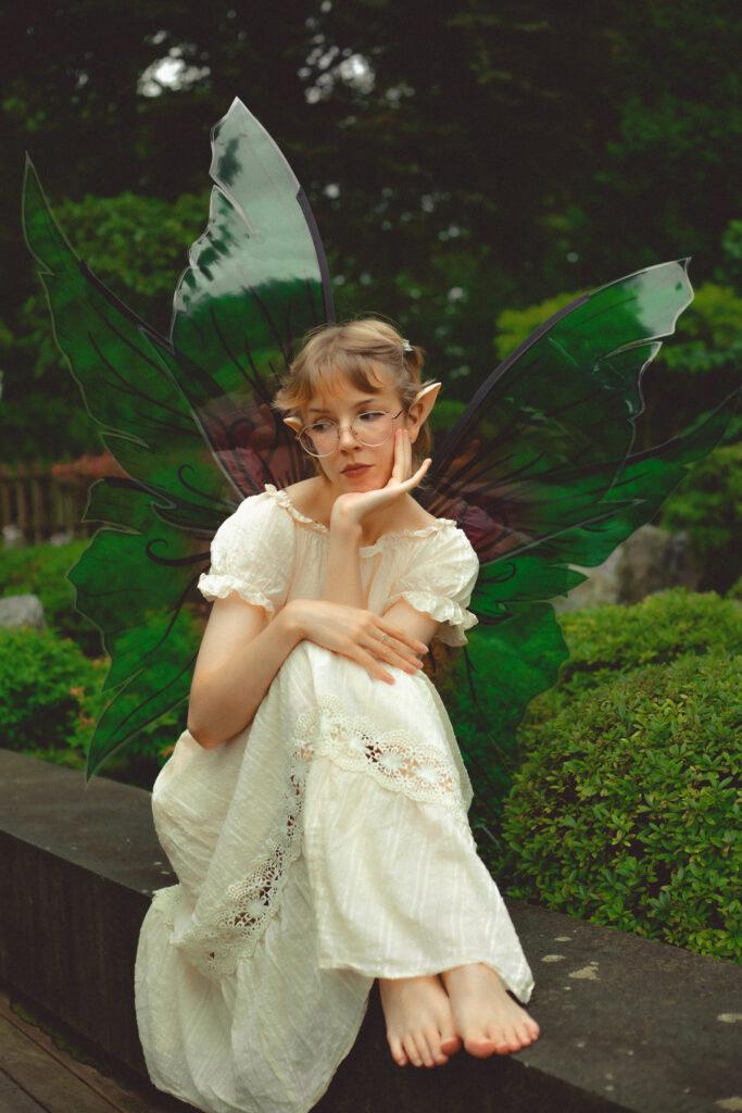 Ein Ganzkörperfoto von einer sitzenden Elfe mit blonden Haaren, hellem Kleid, grün-rose Elfenflügeln und spitzen Ohren.