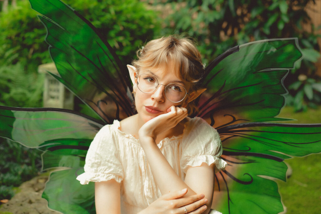 Ein Elfen Portrait eines jungen, blonden Mädchens mit Birlle, spitzen Elfenohren, cremefarbenem Kleid und grün-rosa Flügeln. Im Hintergrund sieht man verschwommen Bäume und Büsche.