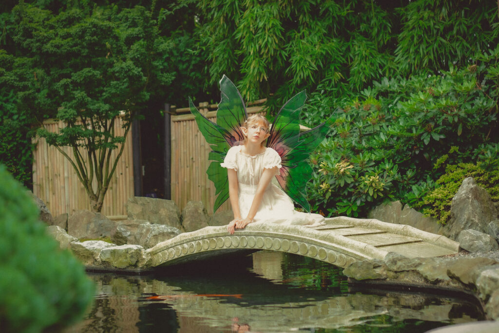 Ein Ganzkörperfoto aus dem Fantasy Fotoshooting der blonden Elfe, mit grün-rosa Flügeln, auf einer Brücke über einem Teich in der Natur.