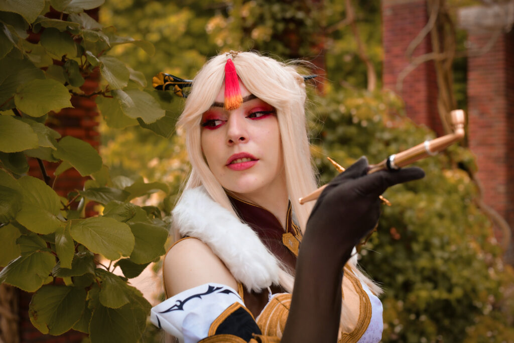 Cosplay Foto der Cosplayerin Alkalashii in ihrem Ningguang Cosplay aus dem Spiel Genshin Impact. Sie steht an einer mit Blättern bewachsenen Säule.