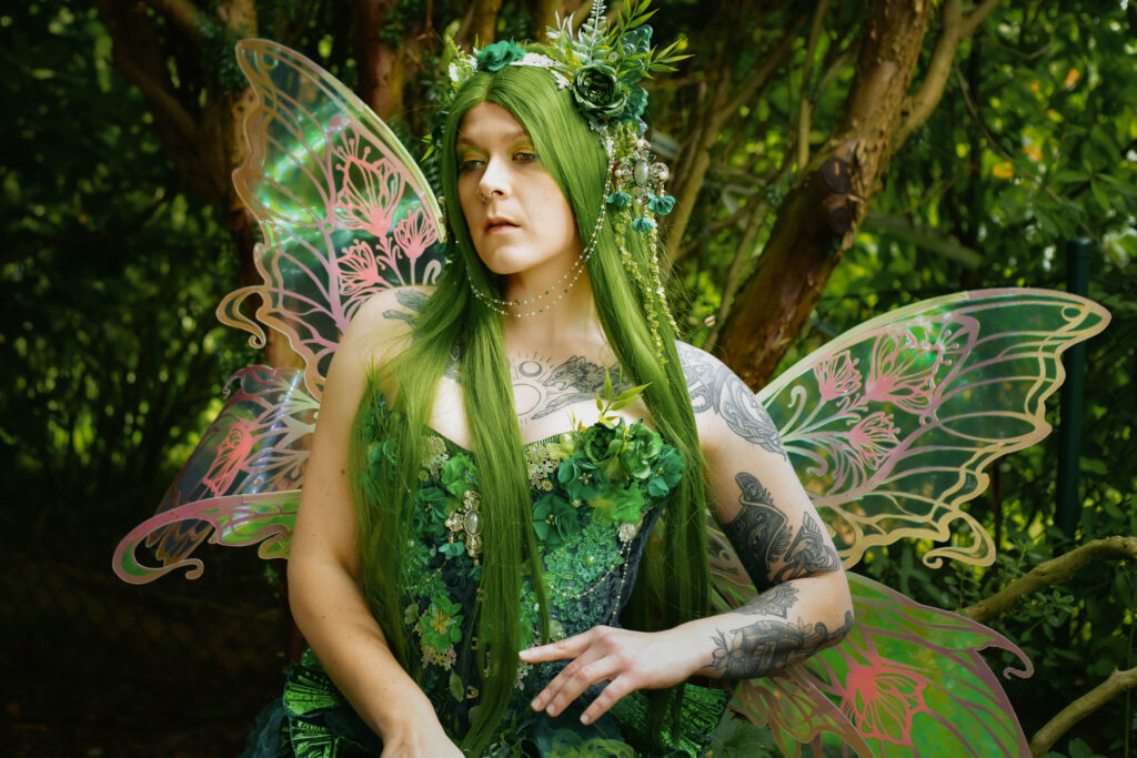 Fantasy Portrait einer Waldelfe mit grünem und Pflanzen überwachsenem Outfit und grünen Flügeln sitzt im Wald auf einer Mauer.