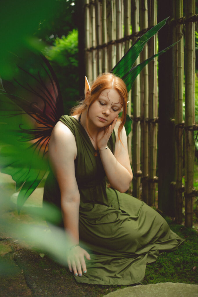 Fantasy Foto einer rothaaringen Elfe im grünen Infinity-Kleid, grün-rosa Flügeln sitzend auf einem Stein inmitten von Moos. Neben ihr ein Bambuszaun.