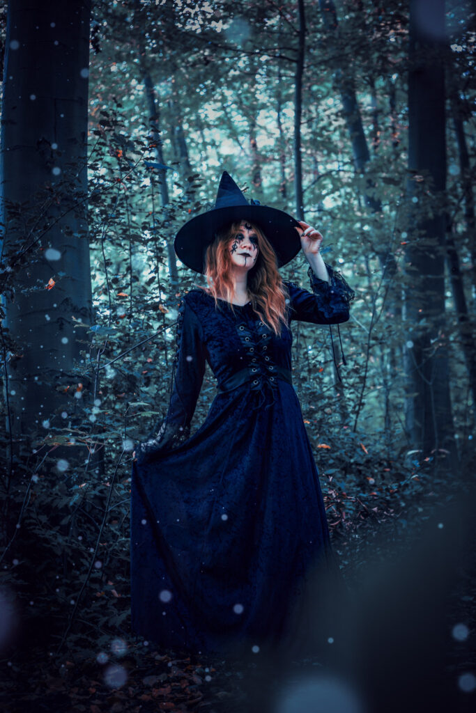 Die Hexe steht im dunklen, verschneiten Wald. Ihr schwarzes langes Kleid, ihr Hut und ihre roten Haaren werden betont.