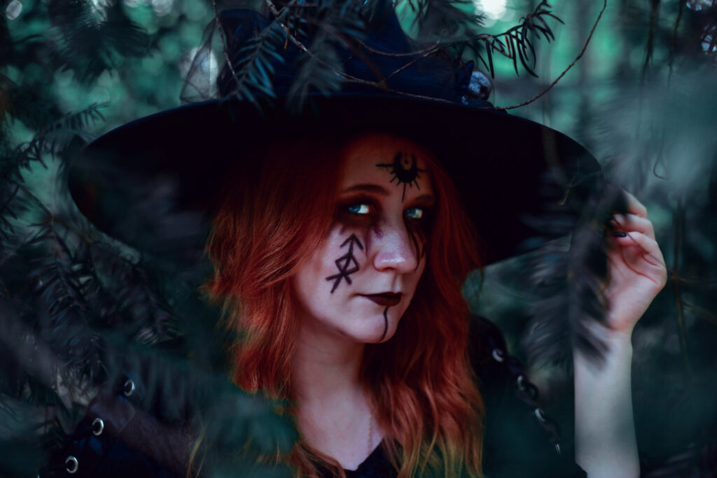 Fantasy Portrait einer Hexe im Wald. Sie hat rote Haare, einen schwarzen Hut und dunkle Glypen und Symbole im Gesicht.