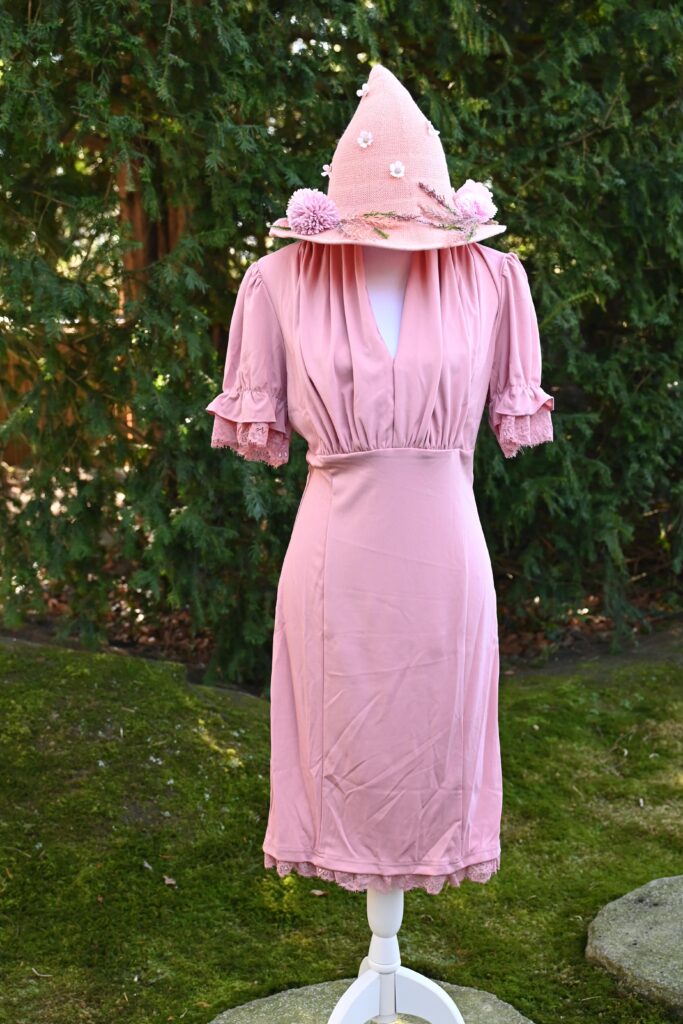 Ein rosa Kleid mit Rüschen auf einer weißen Kleiderpuppe. Oben drauf ein rosa Strickhut mit Blumen.