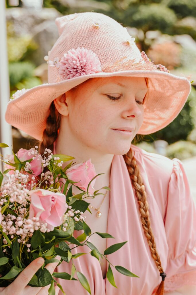 Fantasy-Fotografie einer jungen Frau im rosa Hexenhut mit Zöpfen und einem Strauß rosa Blumen – aufgenommen im Grünen