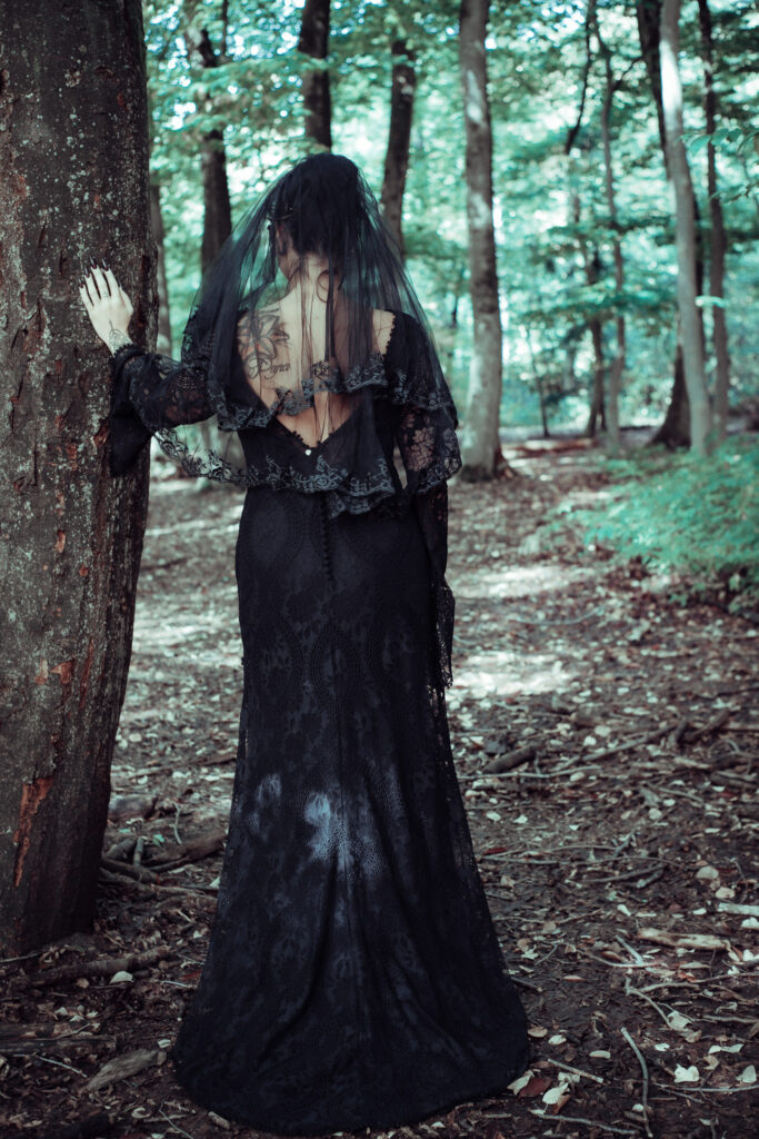 Rückenansicht einer Hexe in schwarzem Kleid und Schleier bei einem mystischen Fantasy-Fotoshooting im Wald.